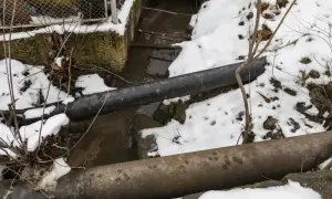 Zamarzanie rur w domku letniskowym. Dwa grube rury biegną nad strumieniem, a wokół leży śnieg.