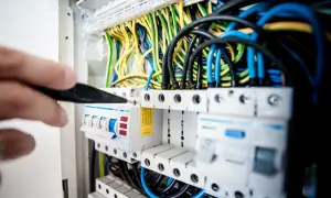 Ręka z narzędziem przyłącze trójfazowe w rozdzielnicy elektrycznej. Widać kolorowe przewody i bezpieczniki.