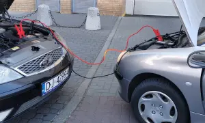 Plus czerwony minus czarny. Dwa samochody podłączone kablami rozruchowymi. Jeden czarny Ford, drugi srebrny Peugeot.
