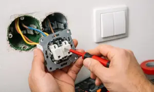 Ręce majsterkowicza podłączają włącznik światła, używając czerwonego śrubokręta. Widać przewody elektryczne i gniazdka w ścianie.