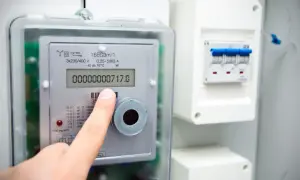 Dłoń wskazuje na wyświetlacz inteligentnego licznika energii elektrycznej, pokazujący zużycie 717.0. Obok widoczny jest wyłącznik.