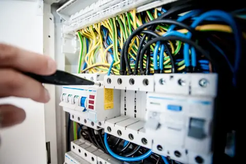 Ręka z narzędziem przyłącze trójfazowe w rozdzielnicy elektrycznej. Widać kolorowe przewody i bezpieczniki.