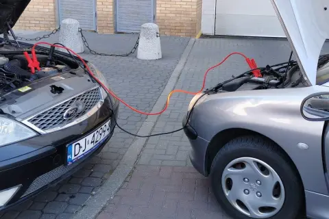 Plus czerwony minus czarny. Dwa samochody podłączone kablami rozruchowymi. Jeden czarny Ford, drugi srebrny Peugeot.