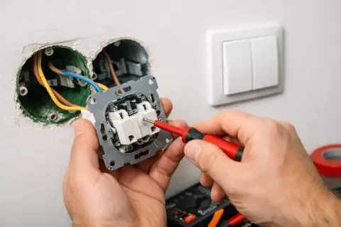 Ręce majsterkowicza podłączają włącznik światła, używając czerwonego śrubokręta. Widać przewody elektryczne i gniazdka w ścianie.