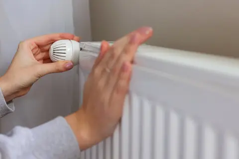 Dłonie regulują termostat grzejnika. Kiedy wyłączyć ogrzewanie w domu, by oszczędzać energię?