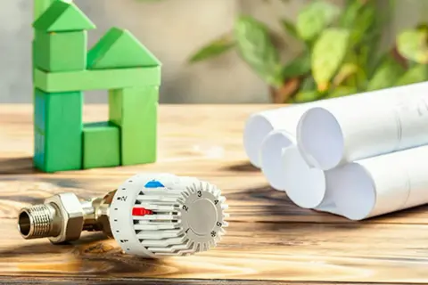 Termostat i zielony domek z klocków symbolizują ogrzewanie ekologiczne rodzaje. Plany domu sugerują projektowanie energooszczędnych rozwiązań.