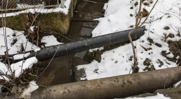 Zamarzanie rur w domku letniskowym. Dwa grube rury biegną nad strumieniem, a wokół leży śnieg.
