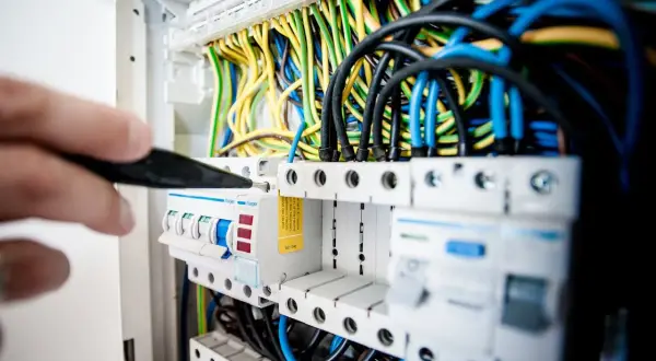 Ręka z narzędziem przyłącze trójfazowe w rozdzielnicy elektrycznej. Widać kolorowe przewody i bezpieczniki.