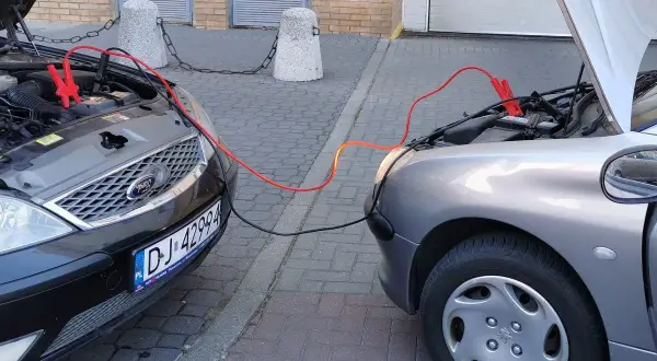 Plus czerwony minus czarny. Dwa samochody podłączone kablami rozruchowymi. Jeden czarny Ford, drugi srebrny Peugeot.