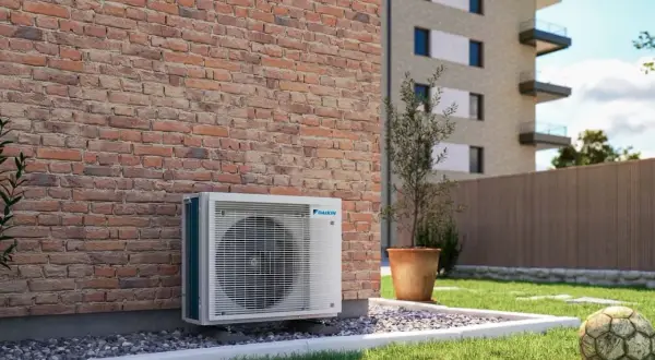 Jednostka zewnętrzna klimatyzatora Daikin przy ceglanej ścianie. Obok trawnik, donica z drzewkiem i stara piłka. Optymalizacja miesięcznego zużycia prądu w mieszkaniu.