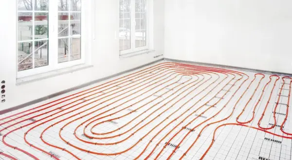 Instalacja ogrzewania podłogowego i grzejników na jednym obiegu. Pomarańczowe rurki ułożone na podłodze w pomieszczeniu z oknami.