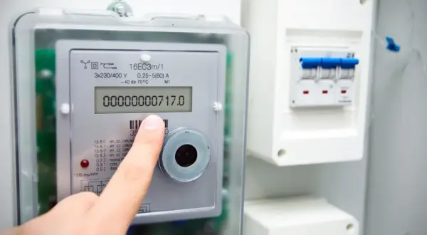 Dłoń wskazuje na wyświetlacz inteligentnego licznika energii elektrycznej, pokazujący zużycie 717.0. Obok widoczny jest wyłącznik.