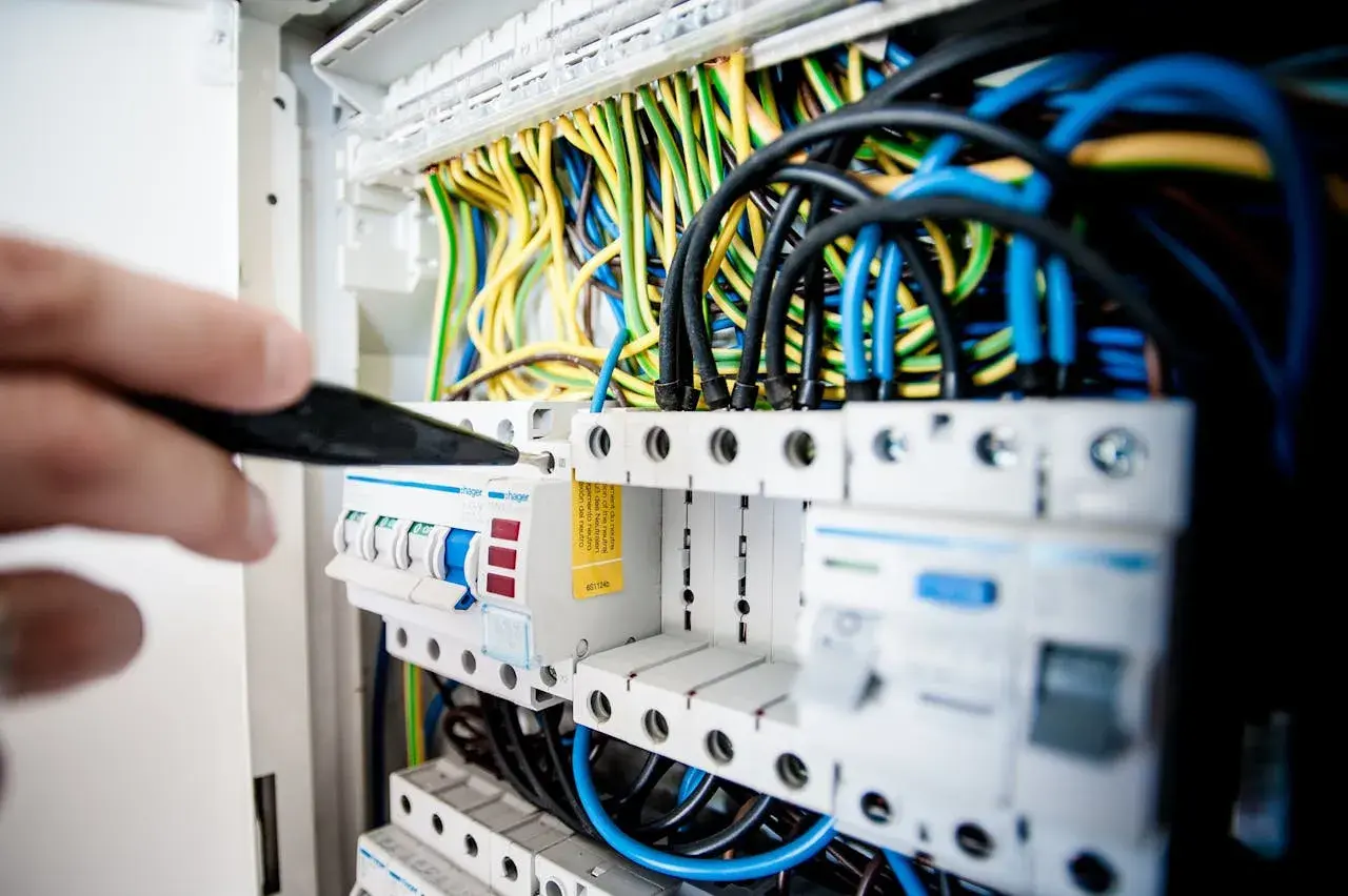 Ręka z narzędziem przyłącze trójfazowe w rozdzielnicy elektrycznej. Widać kolorowe przewody i bezpieczniki.