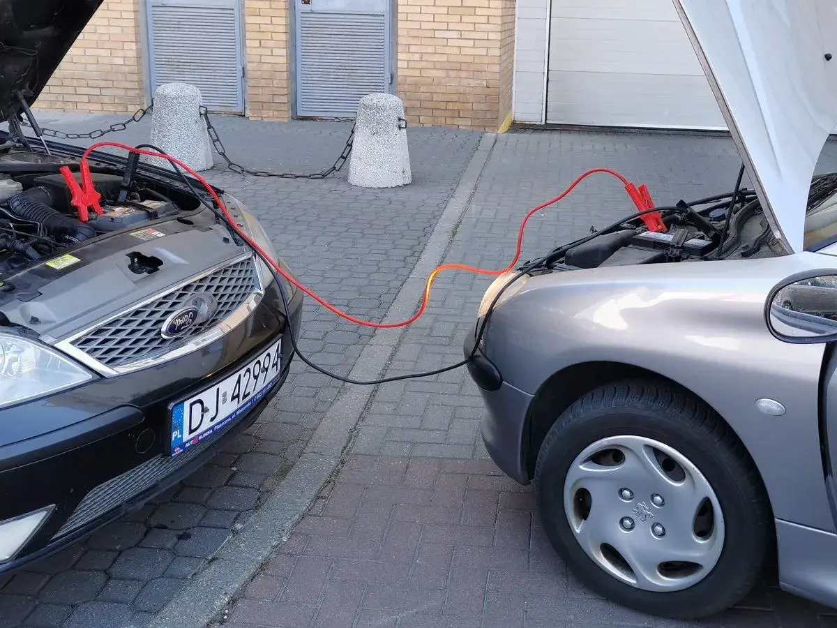 Plus czerwony minus czarny. Dwa samochody podłączone kablami rozruchowymi. Jeden czarny Ford, drugi srebrny Peugeot.