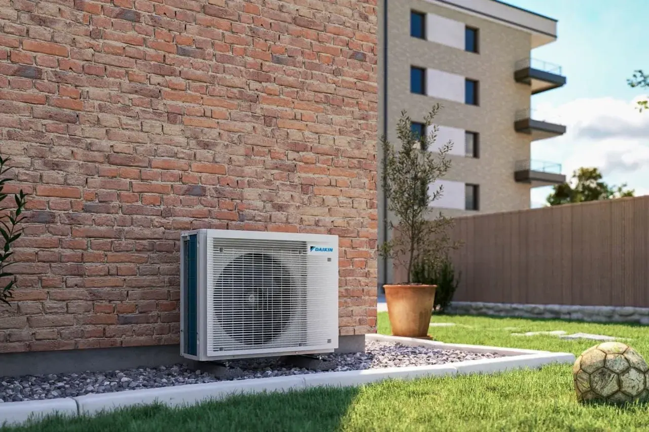 Jednostka zewnętrzna klimatyzatora Daikin przy ceglanej ścianie. Obok trawnik, donica z drzewkiem i stara piłka. Optymalizacja miesięcznego zużycia prądu w mieszkaniu.