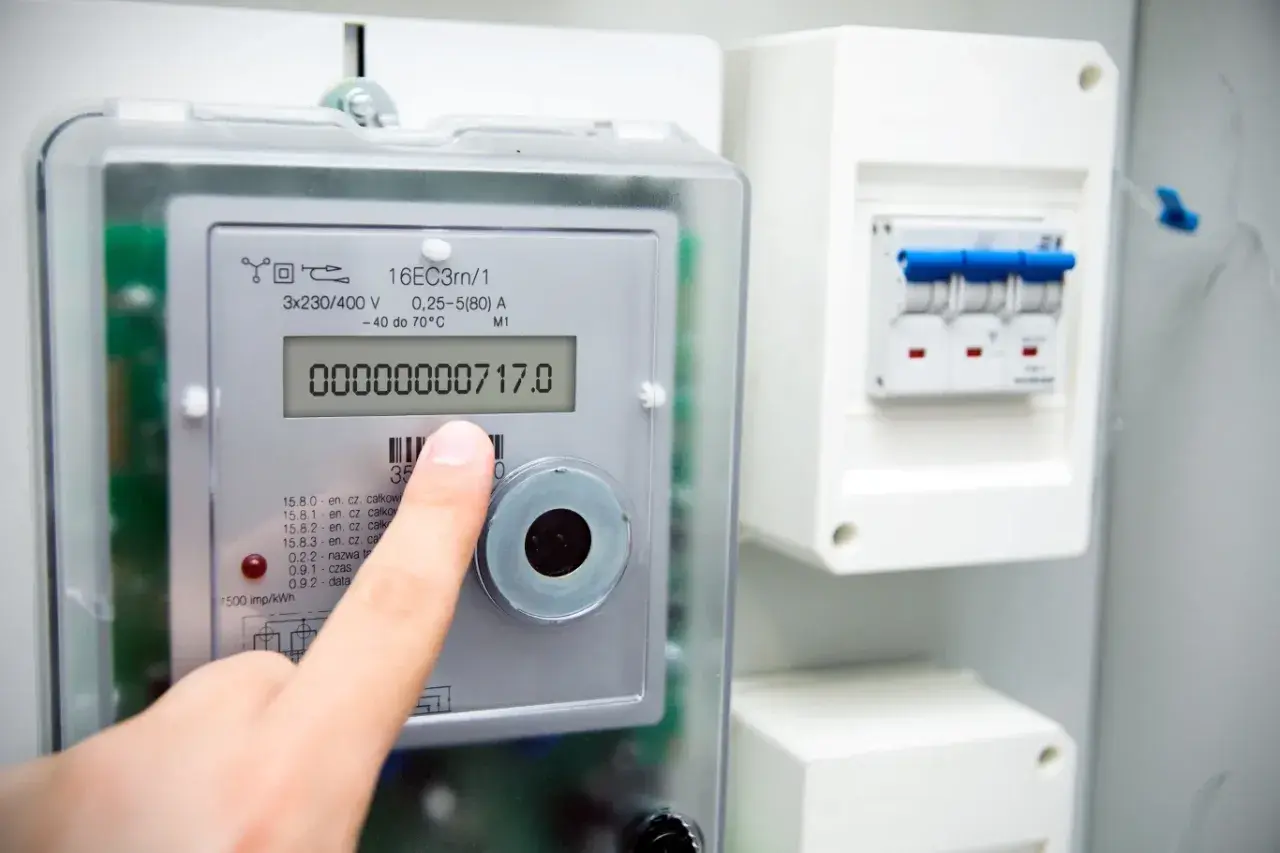 Dłoń wskazuje na wyświetlacz inteligentnego licznika energii elektrycznej, pokazujący zużycie 717.0. Obok widoczny jest wyłącznik.