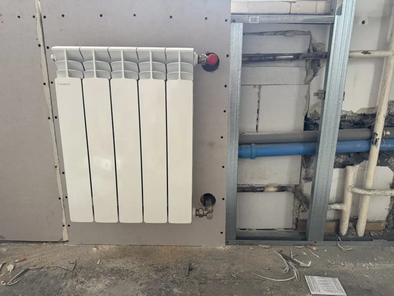 Nowy, biały grzejnik aluminiowy Fondital zamontowany na ścianie z płyt gipsowo-kartonowych. W tle widać instalację hydrauliczną i pustaki. Trwa wymiana grzejnika żeliwnego na aluminiowy.